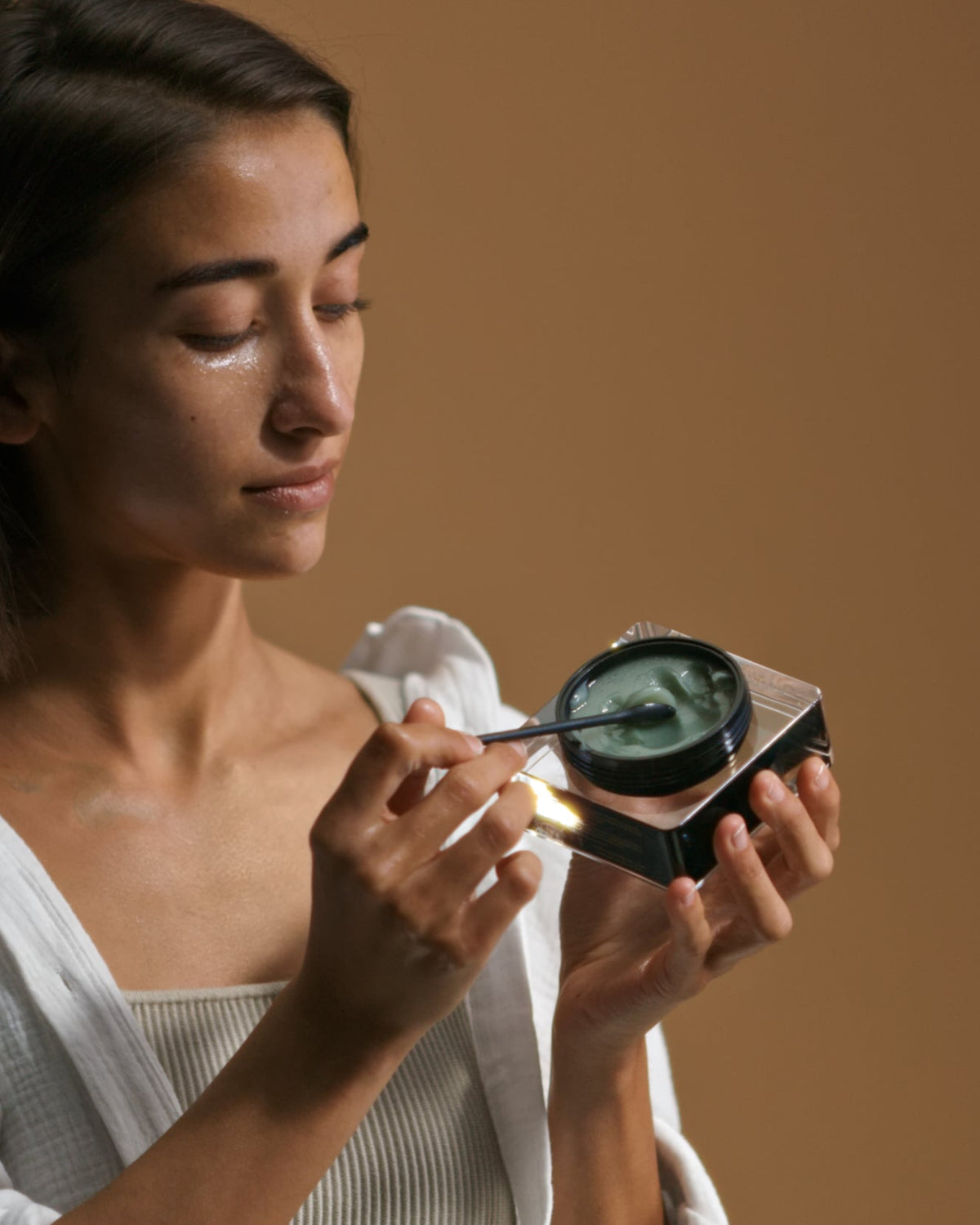 Woman applying cleansing balm to her face with a spatula against a brown background